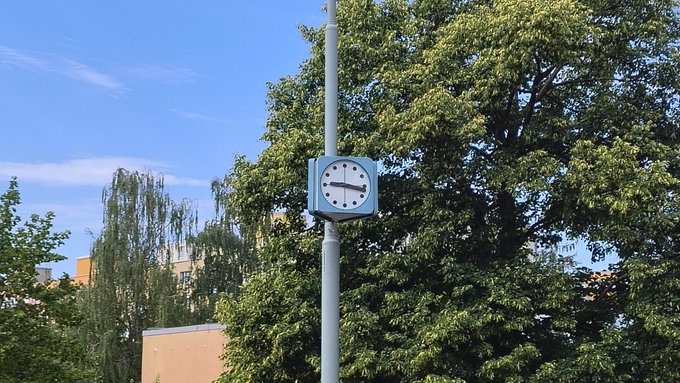 A blue and white analog clock mounted on a pole displays approximately 11:00, with lush green trees and a clear blue sky in the background. The time on the clock matches the post's reference to meeting around 11am at VNITROBLOCK coworking.