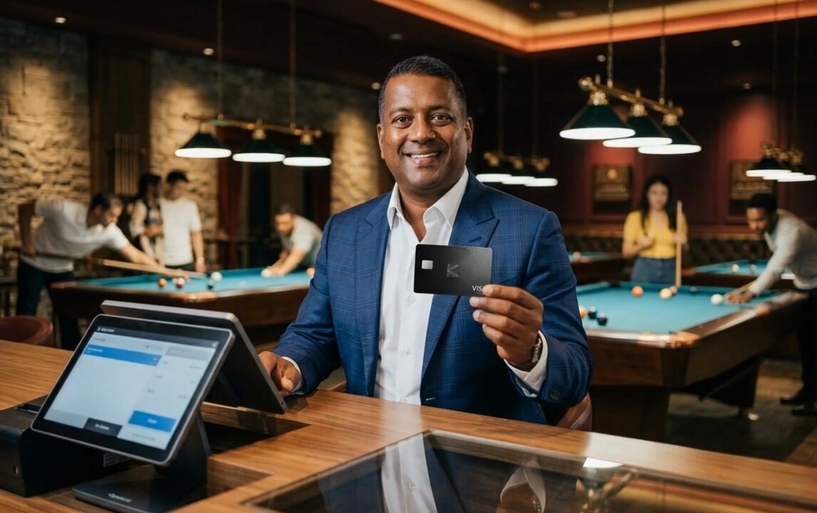 The image features a man sitting at a table with his laptop open, holding up a credit card. The man appears to be smiling as he poses for the camera while showing off his card. In the background, there are other people present who might also be using their laptops or engaging in other activities.