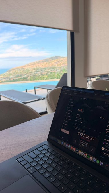 A laptop displaying a crypto portfolio showing $12,224.27 sits on a desk with a stunning ocean and hilltop view in the background, suggesting a remote work or "GM" lifestyle while the owner invites others to drop their wallet addresses in the comments.