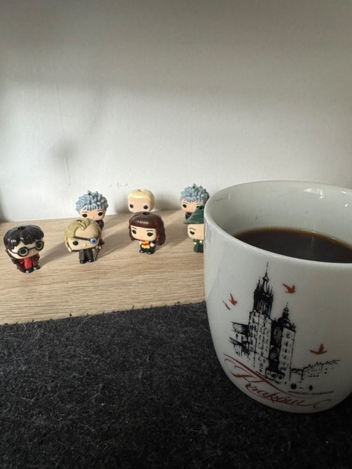 A cozy scene with a white coffee mug labeled 'Prague' featuring a castle illustration and birds, sitting beside a small collection of Funko Pop figurines arranged on a wooden surface.