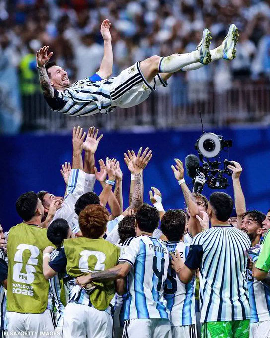 Argentina soccer players in their World Cup 2022 celebration, with Messi being tossed into the air by teammates in blue and white striped jerseys.
