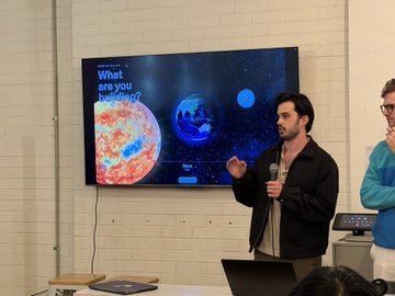 A presenter speaks into a microphone in front of a screen that reads “What are you building?” alongside images of planets, during an OpenAI Devs Build Week session in Melbourne.