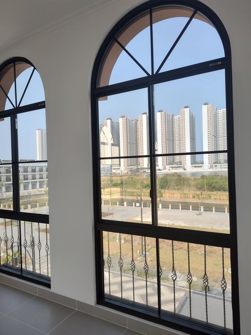 An interior view through two large arched windows with black frames and decorative wrought iron railings, overlooking high-rise apartment buildings and a green field.