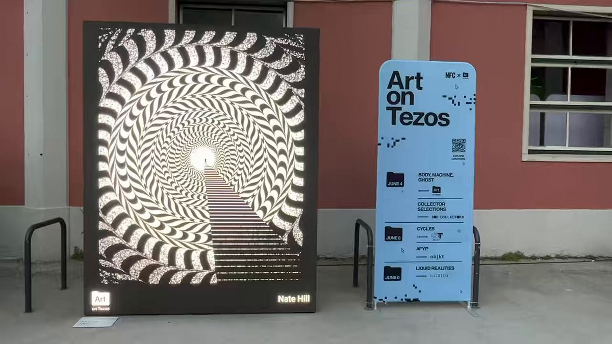 The image features a large poster of a spiral staircase, which is displayed outdoors next to a smaller blue sign that says "Art on Tezos" with some text on it. The poster is positioned near a building, possibly an art gallery or exhibition space. The focus of the image is on these two objects and their arrangement in front of the building.