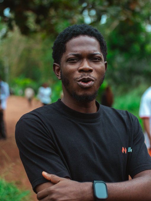 A young Black man with a short beard wears a black t-shirt and a smartwatch, standing outdoors with crossed arms against a backdrop of greenery and blurred figures.