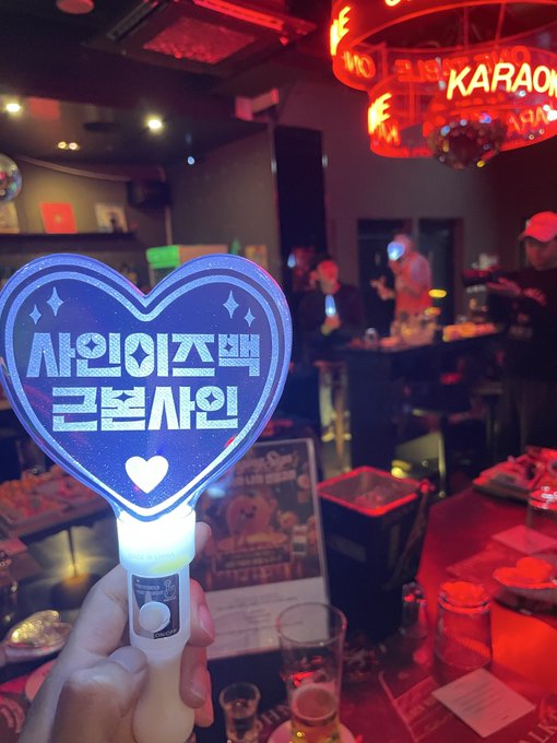 A glowing heart-shaped light stick with Korean characters '사인이즈백 근본시인' held in a dimly lit KARAOKE bar.