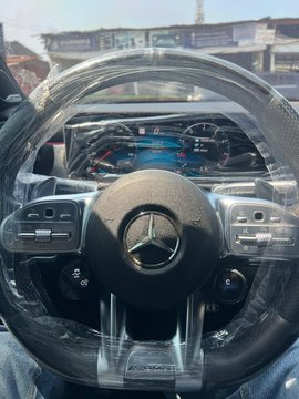 A man sits behind the wheel of his new Mercedes-Benz, the iconic logo prominently displayed on the steering wheel. The dashboard and digital gauges are wrapped in protective plastic, and "AMG" branding is visible below the wheel, capturing the moment of gifting his girlfriend the car to thank her for standing by him through his financial struggles.