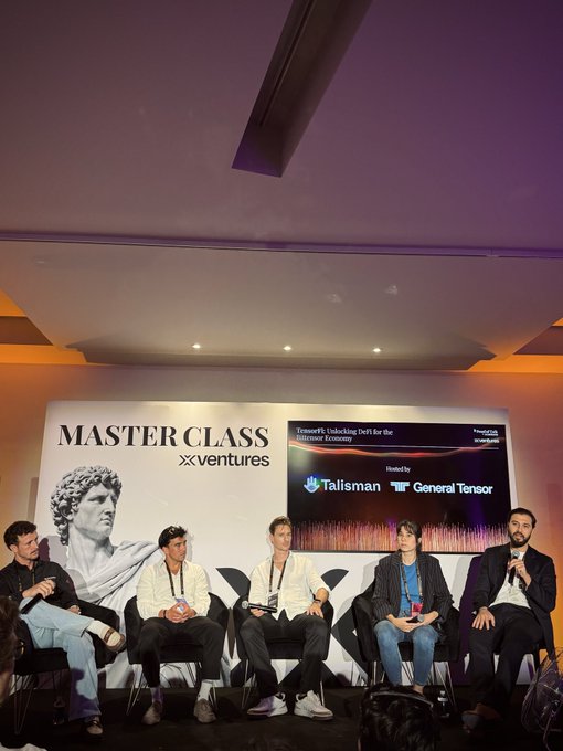The image shows five people sitting around a table, likely participating in a Master Class event or panel discussion. They appear to be engaged in conversation with each other while seated on chairs arranged around the table. The backdrop features a large screen displaying information about the upcoming event and the companies involved.