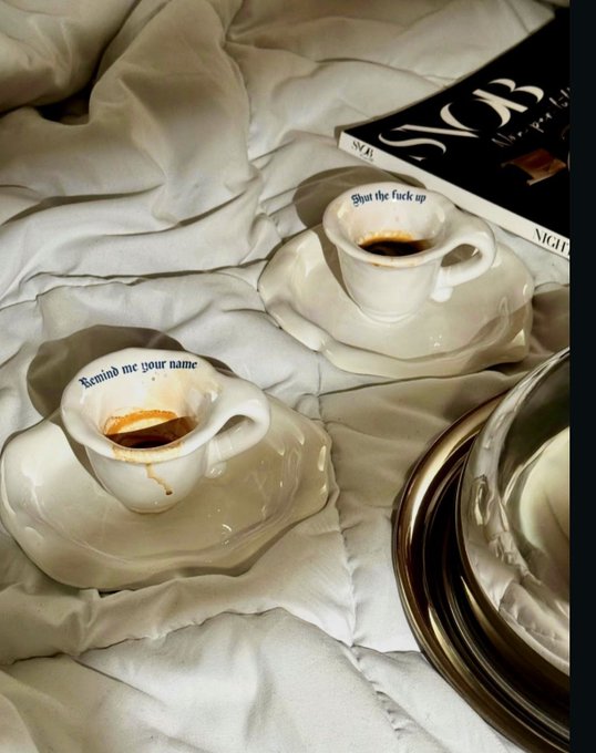 Two small espresso cups on white saucers with saucy text reading 'Remind me your name' and 'Shut the fuck up' placed on a white bed next to a Vogue magazine.