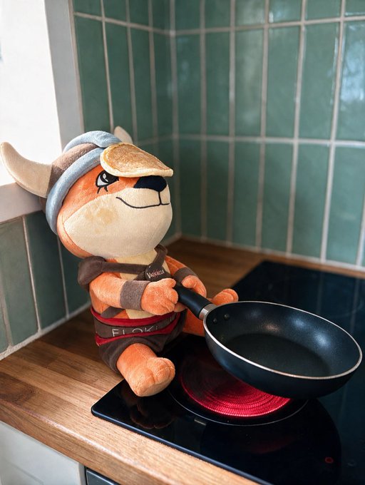 A plush fox mascot wearing a "Flo" apron sits on a stovetop, gripping a black frying pan over a glowing red burner. The charming setup perfectly matches the "Breakfast is served" post, depicting the character cheerfully preparing to cook up a meal.
