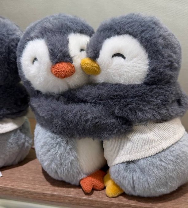 Two plush penguin toys with gray and white fur embrace each other in a hug, featuring orange and yellow beaks and feet.