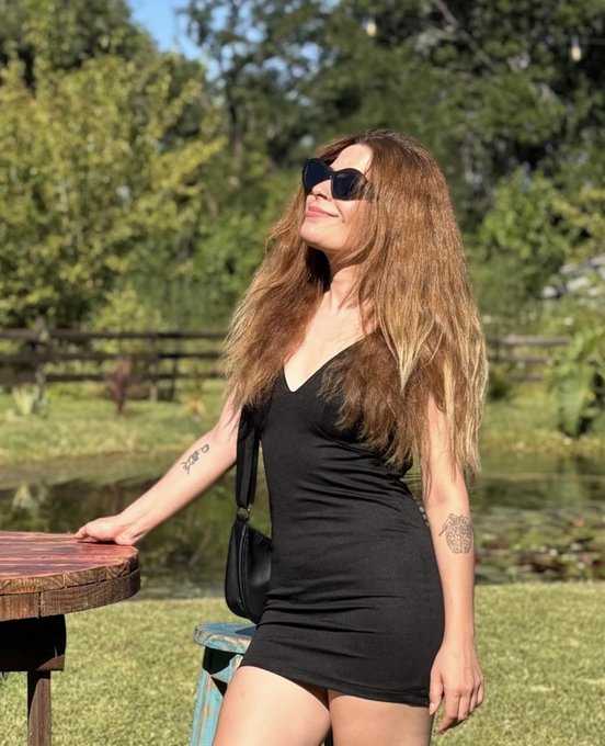 A woman with long brown hair wearing a black dress and sunglasses poses outdoors near a pond, with trees and a fence in the background.