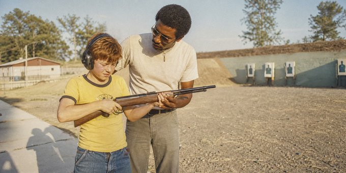 An adult man with sunglasses teaches a young boy in a yellow shirt and ear protection how to hold a shotgun at an outdoor shooting range.