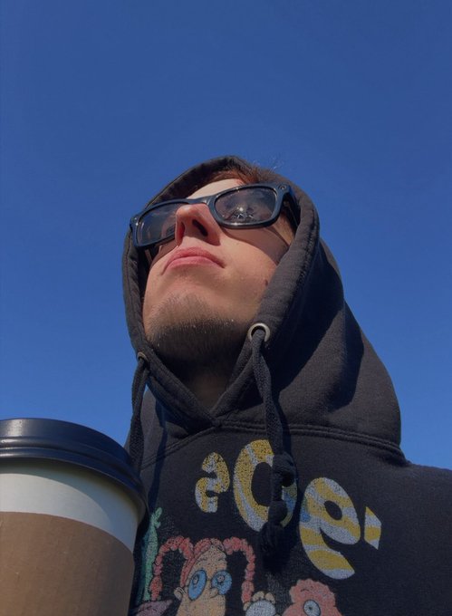 A low-angle selfie of a young man wearing dark-rimmed sunglasses and a black hoodie with colorful graphics, holding a brown coffee cup against a clear blue sky.