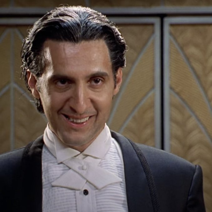 A man with slicked-back dark hair in a formal period costume featuring a white tie and dark jacket, smiling against an ornate gold art deco-style background.