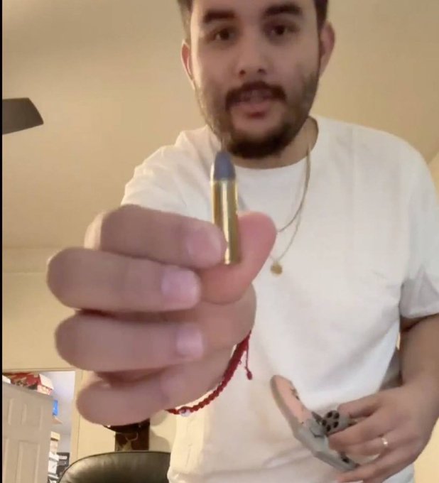 A man in a white shirt holds a small bullet-shaped object close to the camera while also holding a smartphone, wearing a chain necklace and red bracelet.