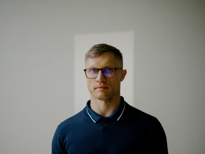 A man wearing a blue sweater and glasses stands against a plain wall with a small window letting in light behind him. The image likely portrays Roman Storm, the Tornado Cash developer whose retrial has been postponed to April 2027 following a mixed verdict in his money-transmitting case.