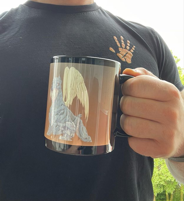 A person wearing a black t-shirt featuring a brown bear paw print logo holds up a black mug decorated with an angel-winged creature, paired with a morning greeting post standing in solidarity with @mutant_cartel on Sunday.