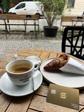 The image features a table with a cup of coffee, a croissant, and a credit card. The coffee cup has a handle and is placed on a saucer along with a small plate holding the croissant. A credit card is also present on the table. In the background, there are chairs and dining tables arranged for people to sit and enjoy their meals or beverages at the café.