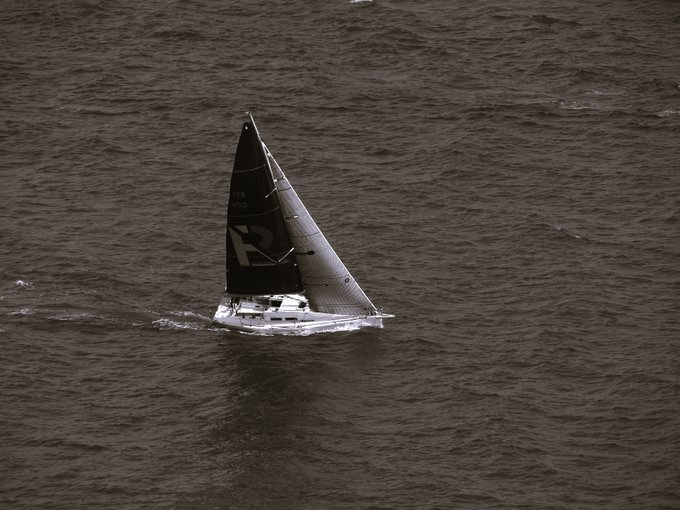 A solitary sailboat with a triangular sail marked by a stylized "R" glides across choppy dark waters, captured in a moody black-and-white composition that emphasizes the vessel's isolation and motion.
