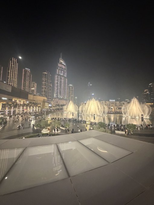 A nighttime cityscape of downtown Dubai featuring the illuminated Burj Khalifa and EMAAR signage, with choreographed fountain displays and crowds gathered around the water. The image visually reinforces the post's bold claim about Dubai's continued dominance in crypto and global events, showcasing the city's iconic skyline as a symbol of resilience and forward momentum.