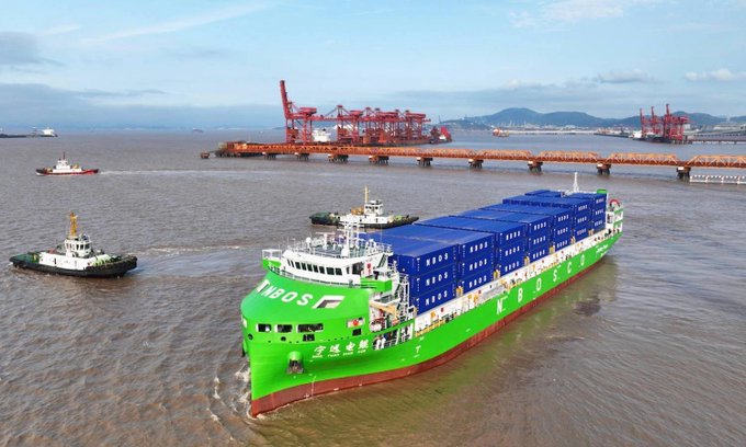 An aerial photograph shows a large green and blue MOSK (Zhejiang Sunway) container ship being escorted by tugboats through a harbor, with red port cranes and a bridge visible in the background.