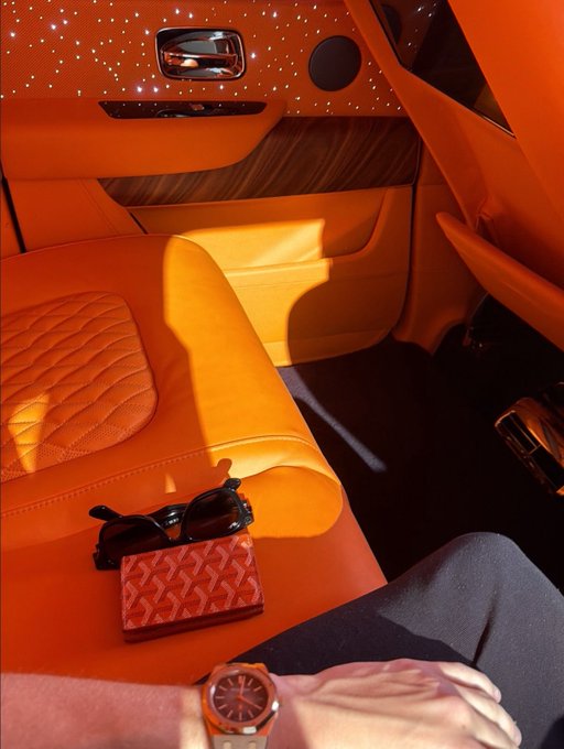 An interior view of a luxury car featuring orange leather seats and a starlight headliner, with sunglasses, a Goyard wallet, and a person's watch-wearing wrist visible in the foreground.