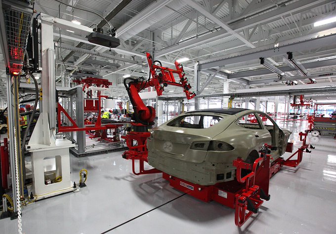 A Tesla car assembly line with a robotic arm and a car body being assembled in a factory.