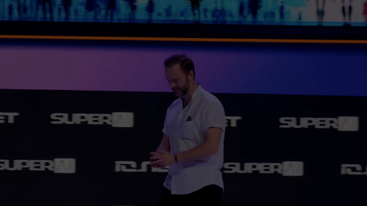 The image features a man standing in front of a large screen or wall with the words "Super" written on it. The man appears to be talking into a microphone, possibly giving a speech or presentation. He is dressed in a white shirt and black pants.