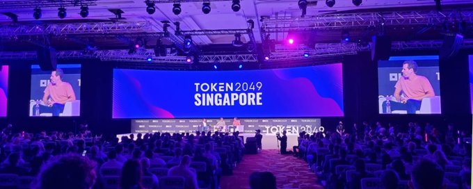 A wide view of a conference hall during the TOKEN2049 Singapore event, featuring a stage with speakers, large screens displaying the event branding, and an audience seated in rows.