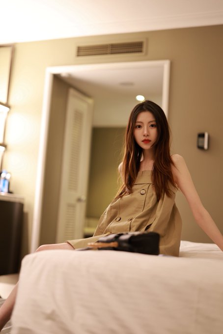 A young woman with long brown hair wearing a beige sleeveless dress sits on the edge of a white bed in a hotel room, looking directly at the camera.