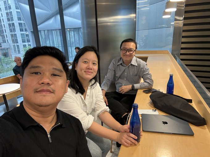 Three people are taking a selfie at a wooden table in a modern café or co-working space, with a man in a black shirt in the foreground, a woman in a white shirt holding a blue bottle, and a man in a grey shirt sitting nearby with a laptop and iPhone.