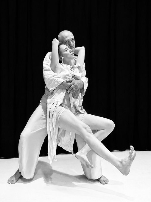 A black-and-white photo of two contemporary dancers in an intimate pose on stage, with the male dancer supporting the female dancer as she leans back with her arms raised.