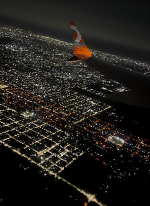 A nighttime aerial view from an airplane window shows the wingtip of a GOL Linhas Aéreas flight, with a vast city grid glowing below as the plane travels to Blockchain Rio.