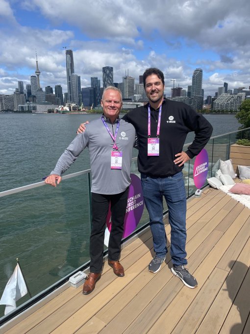 Two smiling men pose together on a wooden deck with a glass railing, overlooking a body of water and the Toronto skyline. The man on the left wears a grey 'T-NODE' zip-up jacket, and the man on the right wears a black 'T-NODE' hoodie; both have lanyards with ID badges, and a pink 'BLOCKCHAIN FUTURES INFINENCE' sign is visible.