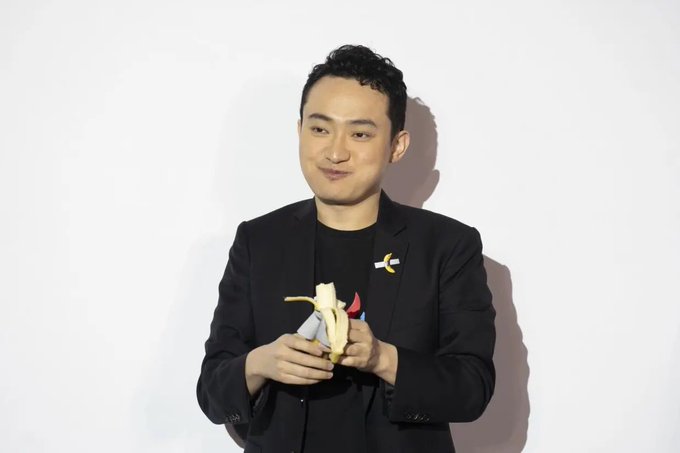 A man in a black suit and t-shirt stands against a white background, peeling a banana and smiling slightly.