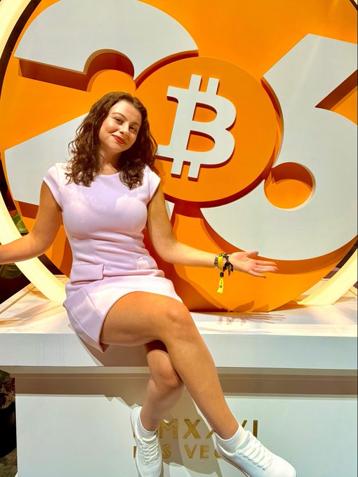 A smiling woman in a light purple dress and white sneakers sits on a platform in front of a large orange Bitcoin logo, with text reading 'MX XII LAS VEGAS' on the base.