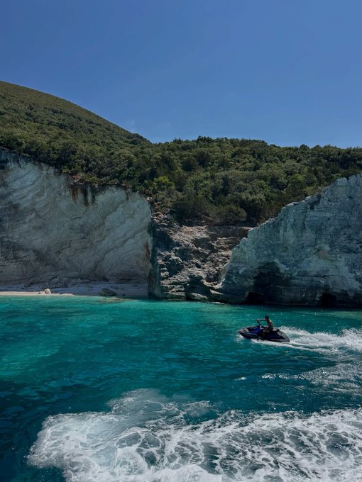 A scenic coastal view featuring turquoise water, white rocky cliffs topped with green vegetation, and a person riding a jet ski.