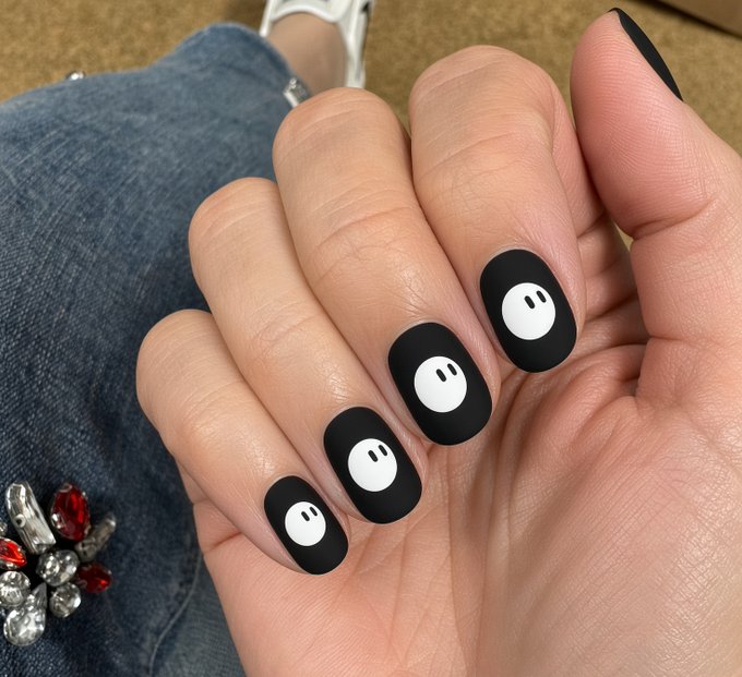 A close-up photo of a hand with black nail polish, each nail featuring a small white cartoon face design, set against denim clothing.