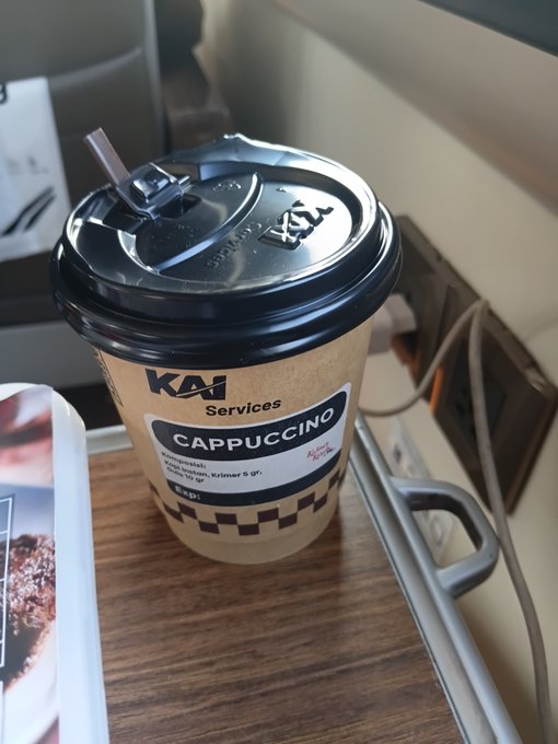 A takeaway paper cup labeled 'KAI Services CAPPUCCINO' with a black plastic lid and brown straw sits on a wooden surface next to a handle.