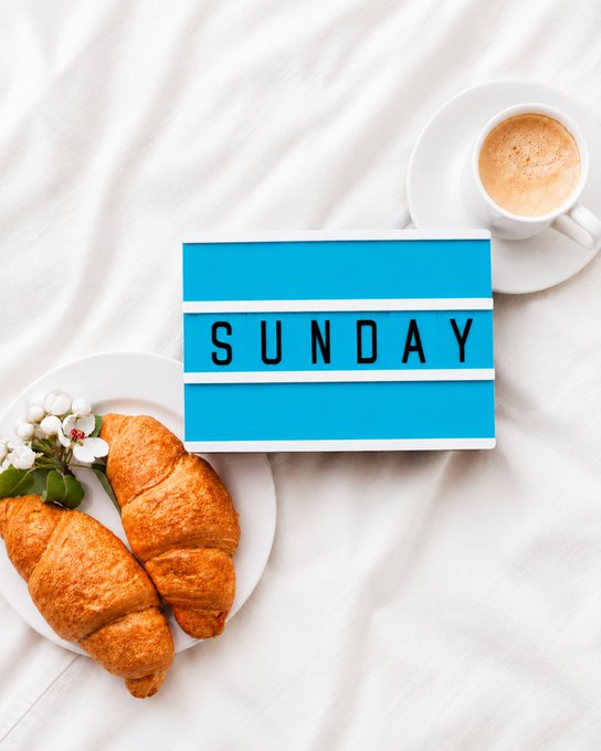 A cozy Sunday morning scene features two golden croissants and a sprig of white berries on a white plate, alongside a cup of frothy coffee, all arranged on soft white bedding. At the center, a lightbox displays "SUNDAY," perfectly capturing the relaxed, leisurely vibe of the "Happy Sunday, folks!" greeting and inviting viewers to share their plans for the day.