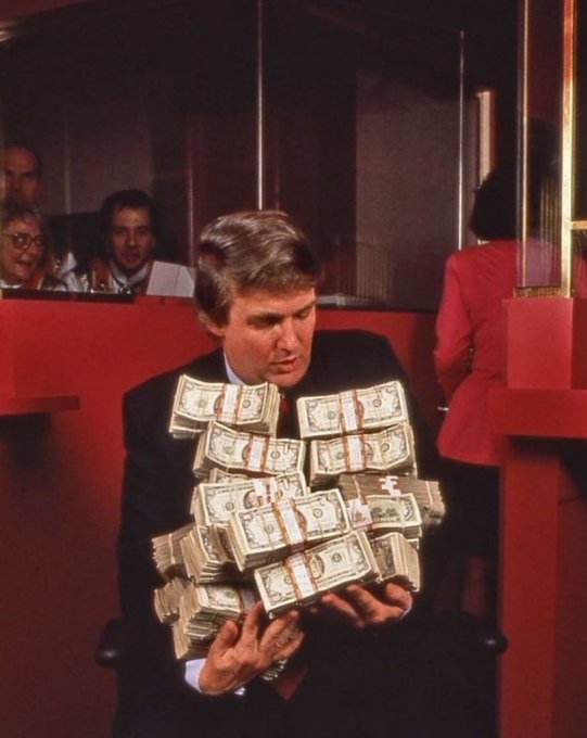 A man in a dark suit holds an enormous stack of bundled US dollar bills, with onlookers visible behind a glass partition.