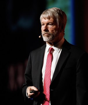 A man with gray hair and a beard, wearing a black suit and red tie, speaks on stage while holding a small device, likely delivering a presentation. The image appears to depict Michael Saylor, founder of Strategy (formerly MicroStrategy, $MSTR), in connection with the news about the company selling 3,588 $BTC worth $225 million.