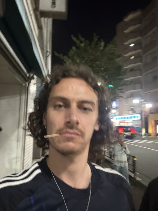 A young man with curly hair takes a nighttime selfie while holding a cigarette in his mouth, with a city street and buildings visible in the background. The post humorously references the old internet practice of "A/S/L" (age/sex/location), suggesting he's playfully sharing that information through the photo.