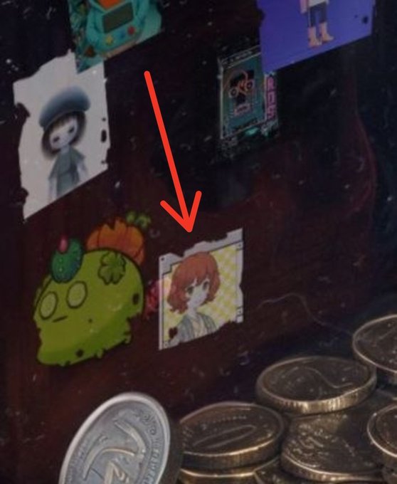 A close-up of a dark surface with various cartoon stickers and coins scattered about, with a red arrow pointing to a sticker of an anime-style character with red hair.
