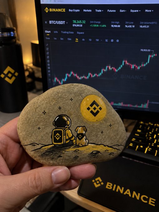 A hand holds a painted stone depicting an astronaut and dog gazing at a Binance logo moon, set against a laptop showing the BTC/USDT price at $78,245.22 on the Binance exchange. The image ties into the "single small step" theme by symbolizing the early, hopeful journey of a crypto investor reaching for Bitcoin's lunar highs.