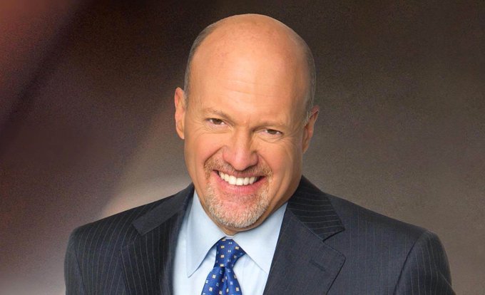 A professional headshot of a smiling bald man with a goatee, wearing a dark pinstripe suit, light blue shirt, and blue patterned tie, set against a brown background.