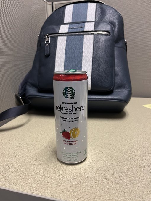 A can of Starbucks Refreshers Strawberry Lemonade sparkling juice drink sitting on a counter in front of a navy blue Coach backpack.