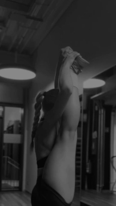 A black-and-white photograph shows a person with a braided ponytail stretching their arms overhead in a gym setting.