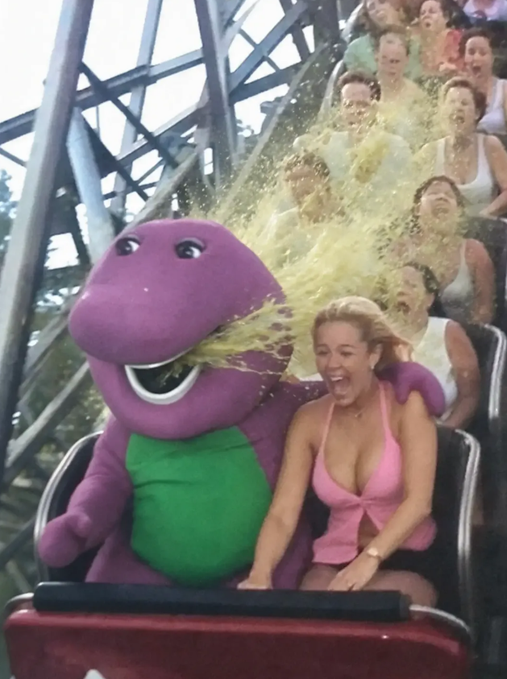 People riding a roller coaster, with a woman in a pink bikini top being splashed in the face with yellow liquid next to Barney the purple dinosaur mascot.